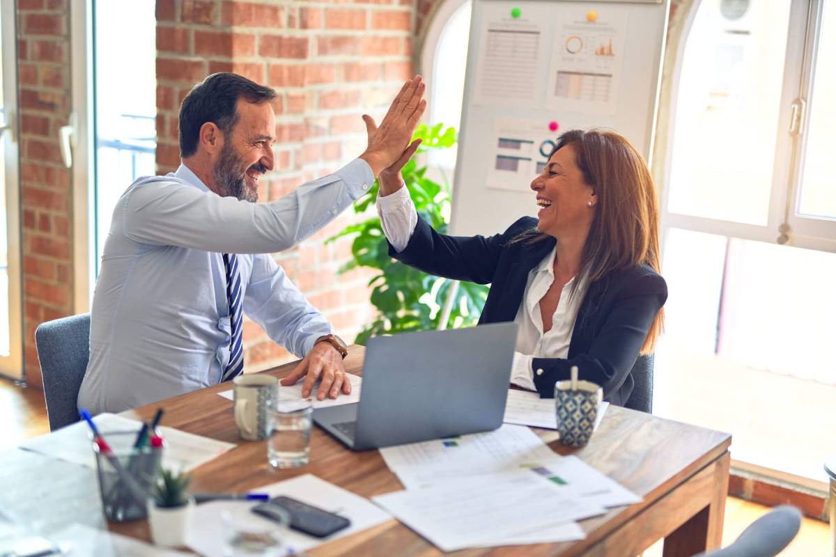 Two colleagues working at a desk next to each other with a laptop and giving each other a high-five